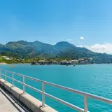 Un muelle con barandilla metálica conduce a un pueblo sobre el agua con montañas verdes de fondo. Ideal para excursiones en Puerto Plata.