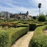 Jardín geométrico con setos cuidados y flores en Villafranca del Bierzo. Al fondo, edificios históricos y una palmera solitaria bajo un cielo azul.