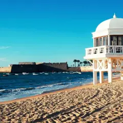 Edificio blanco sobre pilotes en una playa de arena, con el mar azul y un fuerte histórico al fondo. Un ejemplo de las mejores playas de Andalucía.