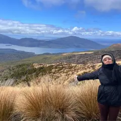 Mujer con los brazos extendidos en el Tongariro Alpine Crossing, disfrutando del paisaje montañoso y el lago azul bajo un cielo nublado.