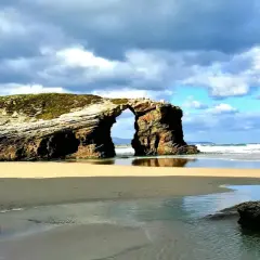 Arco natural en una playa de arena dorada, una de las mejores playas de Galicia, con el mar y el cielo nublado de fondo.
