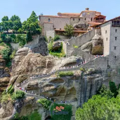 Escaleras de piedra serpentean hacia los impresionantes monasterios de Meteora, construidos en acantilados rocosos. Turistas exploran este sitio histórico.