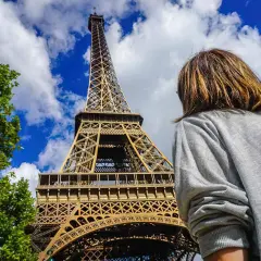 Persona mira la Torre Eiffel, preguntándose cuanto cuesta subir a la torre Eiffel. El cielo azul y las nubes completan la escena.