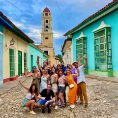 Grupo de amigos sonríe y saluda en una calle empedrada de Trinidad, Cuba, con casas coloridas y una torre de iglesia al fondo. ¡Es seguro viajar a Cuba y disfrutar de su encanto!