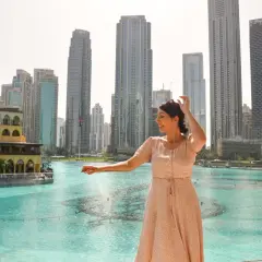 Mujer sonríe mientras disfruta de su viaje a Dubai, con rascacielos y una fuente de agua de fondo.