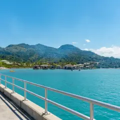 Un muelle con barandilla metálica conduce a un pueblo sobre el agua con montañas verdes de fondo. Ideal para excursiones en Puerto Plata.