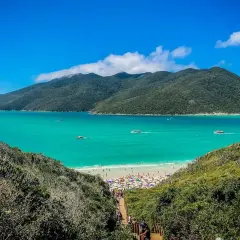 Escenario idílico para viajar a Brasil: playa con aguas turquesas, montañas verdes y gente disfrutando bajo sombrillas.