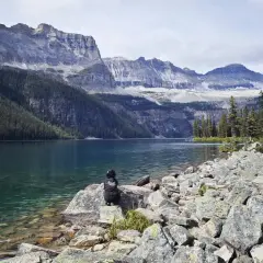 Persona sentada en rocas junto a un lago turquesa, contemplando majestuosas montañas nevadas. Un paisaje perfecto para viajar a Canadá.