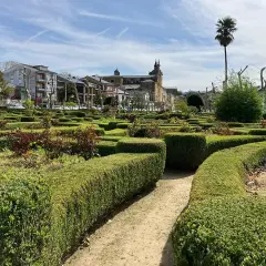 Jardín geométrico con setos cuidados y flores en Villafranca del Bierzo. Al fondo, edificios históricos y una palmera solitaria bajo un cielo azul.