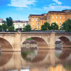 Puente de piedra sobre el río Ebro, reflejándose en sus aguas. Edificios amarillos y verdes árboles componen el paisaje de **que ver en Logroño**.