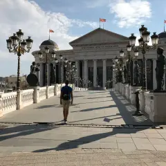 Un hombre camina por un puente adornado con estatuas y farolas, frente al Museo Arqueológico de Macedonia. ¡Un lugar imperdible que ver en Skopje!