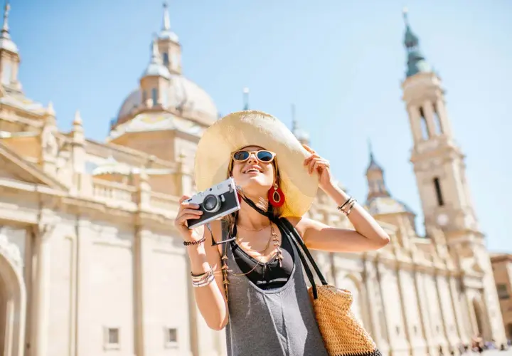 Mujer con sombrero y cámara, lista para viajar por España, con la Basílica del Pilar de fondo.