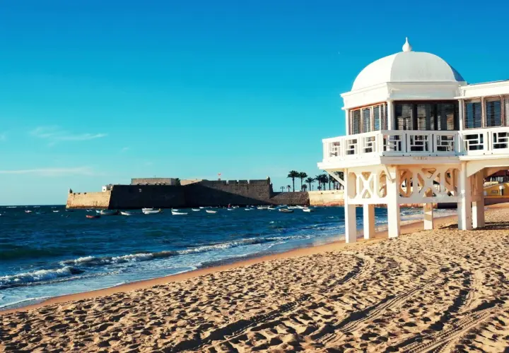 Edificio blanco sobre pilotes en una playa de arena, con el mar azul y un fuerte histórico al fondo. Un ejemplo de las mejores playas de Andalucía.