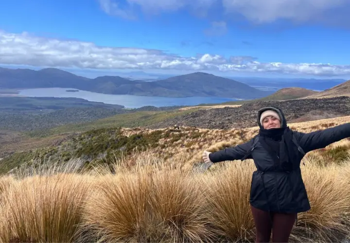 Mujer con los brazos extendidos en el Tongariro Alpine Crossing, disfrutando del paisaje montañoso y el lago azul bajo un cielo nublado.
