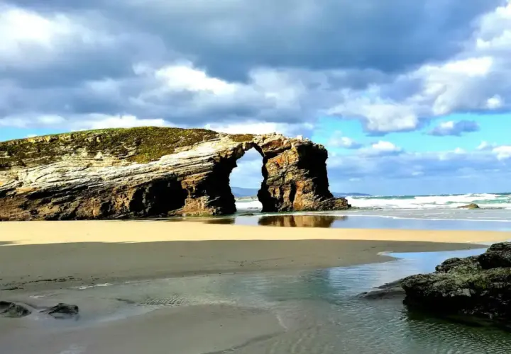 Arco natural en una playa de arena dorada, una de las mejores playas de Galicia, con el mar y el cielo nublado de fondo.