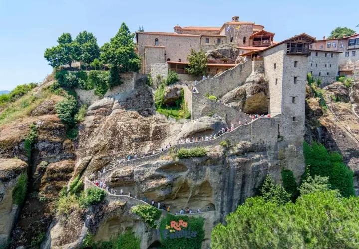 Escaleras de piedra serpentean hacia los impresionantes monasterios de Meteora, construidos en acantilados rocosos. Turistas exploran este sitio histórico.