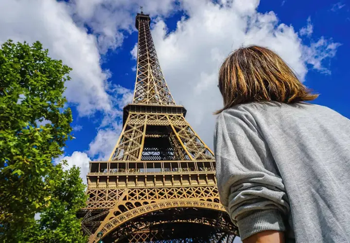 Persona mira la Torre Eiffel, preguntándose cuanto cuesta subir a la torre Eiffel. El cielo azul y las nubes completan la escena.