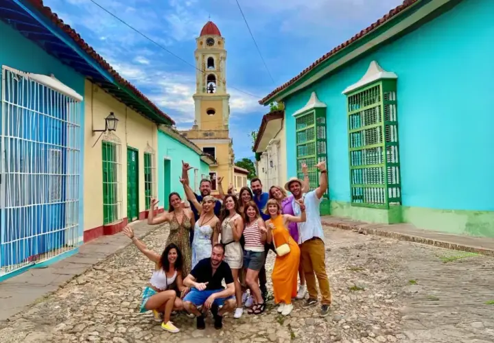 Grupo de amigos sonríe y saluda en una calle empedrada de Trinidad, Cuba, con casas coloridas y una torre de iglesia al fondo. ¡Es seguro viajar a Cuba y disfrutar de su encanto!