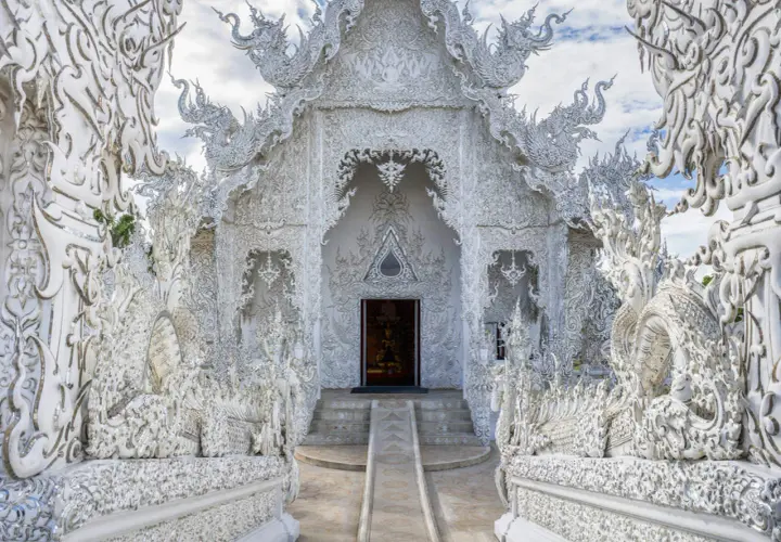 El Wat Rong Khun, un templo blanco deslumbrante en Tailandia, exhibe intrincadas esculturas de dragones y motivos ornamentados bajo un cielo azul.