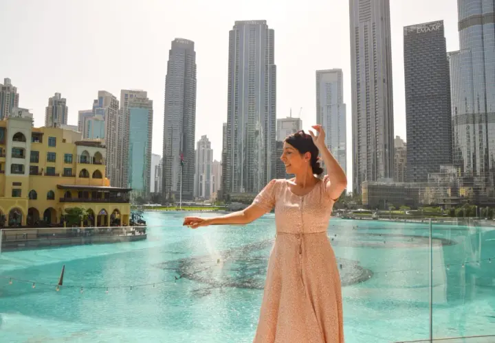 Mujer sonríe mientras disfruta de su viaje a Dubai, con rascacielos y una fuente de agua de fondo.