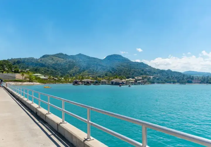 Un muelle con barandilla metálica conduce a un pueblo sobre el agua con montañas verdes de fondo. Ideal para excursiones en Puerto Plata.