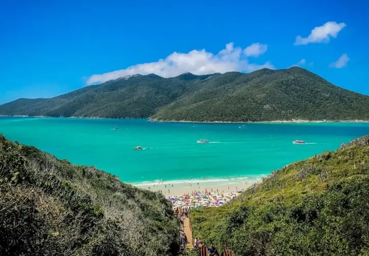 Escenario idílico para viajar a Brasil: playa con aguas turquesas, montañas verdes y gente disfrutando bajo sombrillas.