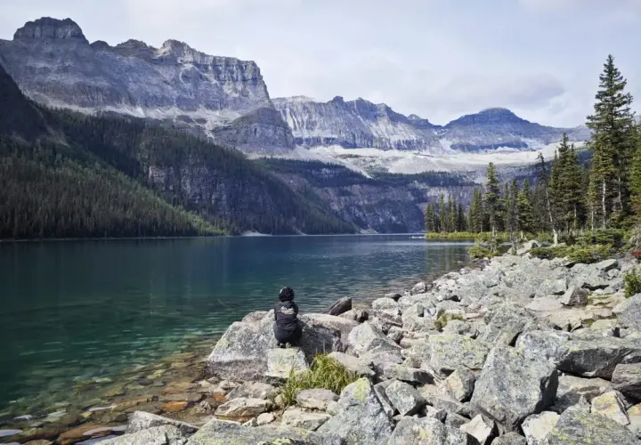 Persona sentada en rocas junto a un lago turquesa, contemplando majestuosas montañas nevadas. Un paisaje perfecto para viajar a Canadá.