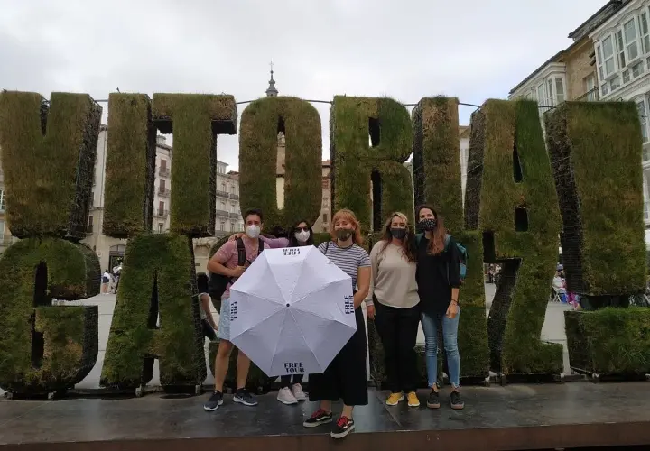 Grupo posa frente a letras gigantes de césped que dicen "VITORIA". Sostienen un paraguas blanco con el logo "FREE TOUR".