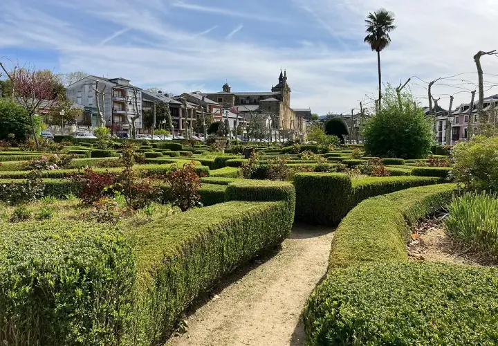 Jardín geométrico con setos cuidados y flores en Villafranca del Bierzo. Al fondo, edificios históricos y una palmera solitaria bajo un cielo azul.