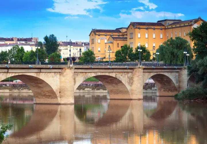 Puente de piedra sobre el río Ebro, reflejándose en sus aguas. Edificios amarillos y verdes árboles componen el paisaje de **que ver en Logroño**.