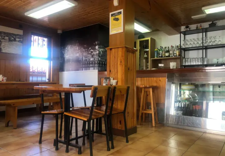 Mesas de madera y sillas listas para comer en Pontedeume. Barra con botellas y vasos, y un mural de la ciudad.