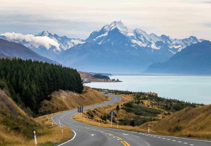 Carretera serpenteante hacia montañas nevadas y un lago turquesa. Impresionantes vistas de Nueva Zelanda que ver.