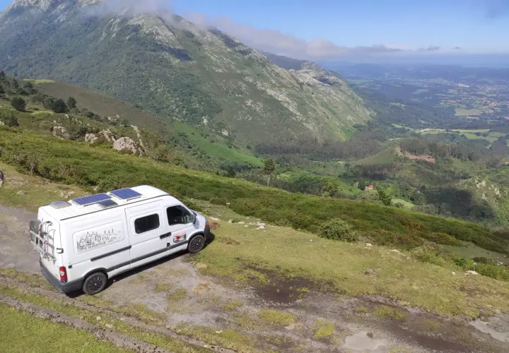 Furgoneta blanca con paneles solares aparcada en una ladera verde, lista para viajar en furgoneta y explorar las montañas.