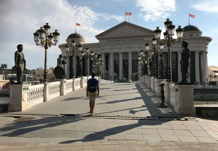 Un hombre camina por un puente adornado con estatuas y farolas, frente al Museo Arqueológico de Macedonia. ¡Un lugar imperdible que ver en Skopje!