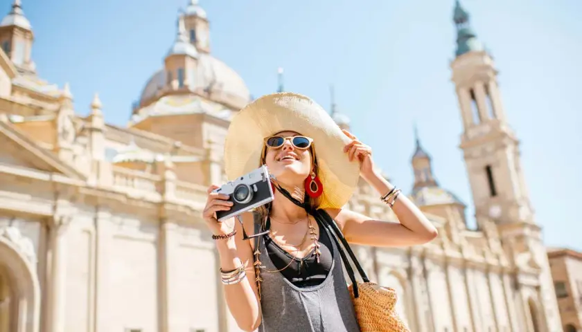 Mujer con sombrero y cámara, lista para viajar por España, con la Basílica del Pilar de fondo.