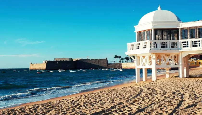 Edificio blanco sobre pilotes en una playa de arena, con el mar azul y un fuerte histórico al fondo. Un ejemplo de las mejores playas de Andalucía.