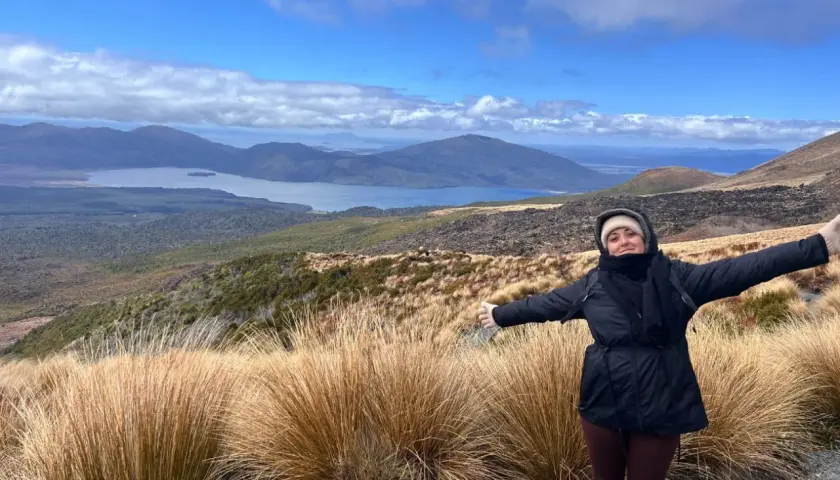 Mujer con los brazos extendidos en el Tongariro Alpine Crossing, disfrutando del paisaje montañoso y el lago azul bajo un cielo nublado.