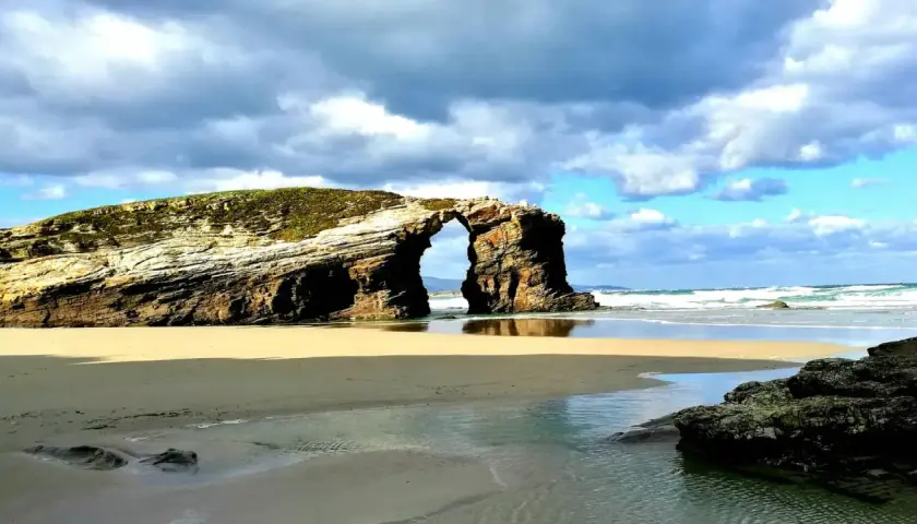Arco natural en una playa de arena dorada, una de las mejores playas de Galicia, con el mar y el cielo nublado de fondo.