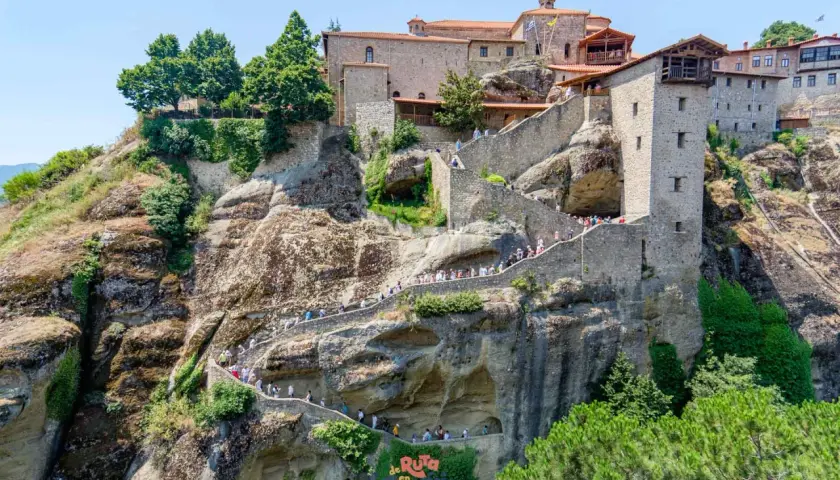 Escaleras de piedra serpentean hacia los impresionantes monasterios de Meteora, construidos en acantilados rocosos. Turistas exploran este sitio histórico.