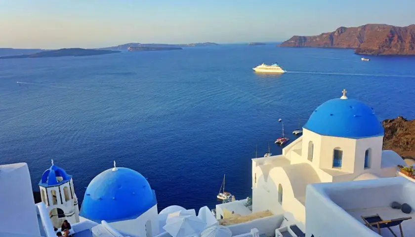 Iglesias blancas con cúpulas azules y el mar Egeo azul profundo. Un crucero navega en la distancia. ¡Imprescindible que ver en Santorini!