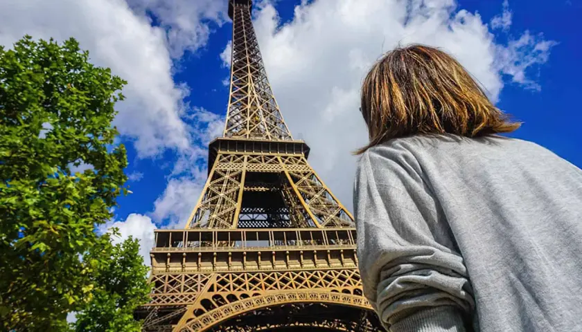 Persona mira la Torre Eiffel, preguntándose cuanto cuesta subir a la torre Eiffel. El cielo azul y las nubes completan la escena.