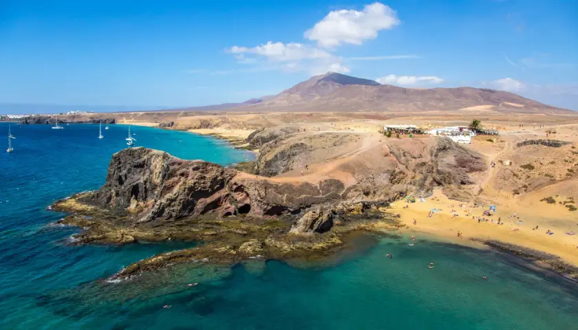 Costa de Lanzarote con playas doradas, aguas turquesas y un restaurante en la colina. ¡Perfecto para tu plan de Lanzarote en 4 días!
