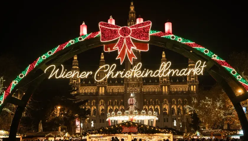Un mercado navideño en Viena, con un arco de luces y un gran lazo rojo. El edificio del ayuntamiento ilumina el fondo.