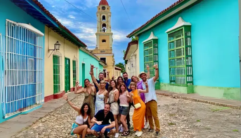 Grupo de amigos sonríe y saluda en una calle empedrada de Trinidad, Cuba, con casas coloridas y una torre de iglesia al fondo. ¡Es seguro viajar a Cuba y disfrutar de su encanto!