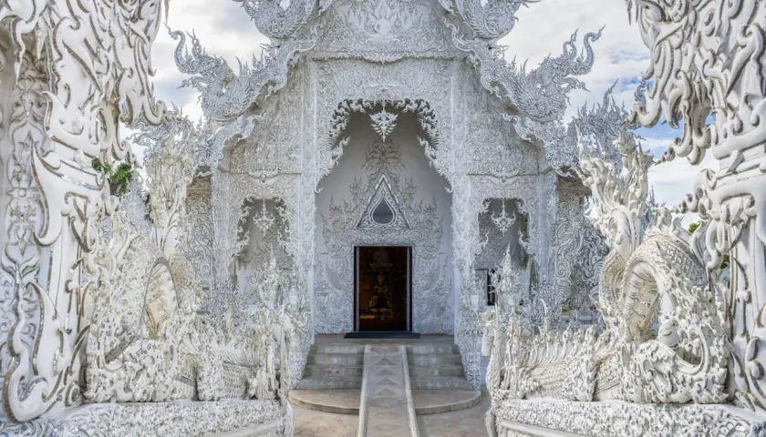 El Wat Rong Khun, un templo blanco deslumbrante en Tailandia, exhibe intrincadas esculturas de dragones y motivos ornamentados bajo un cielo azul.