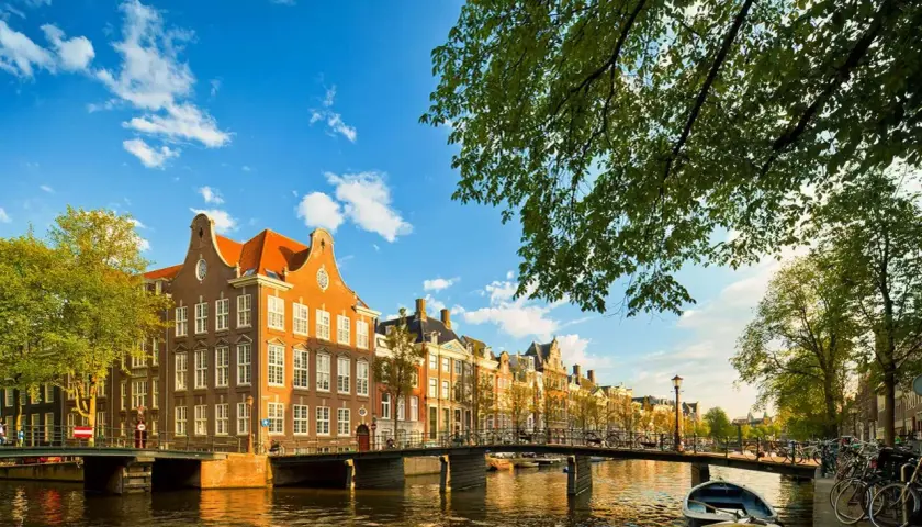 Un día perfecto para viajar a Ámsterdam: casas históricas junto a un canal, puentes y barcas bajo un cielo azul.