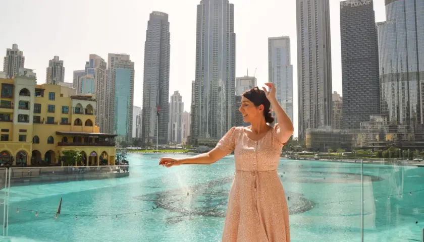 Mujer sonríe mientras disfruta de su viaje a Dubai, con rascacielos y una fuente de agua de fondo.