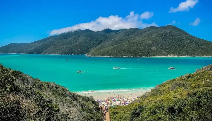 Escenario idílico para viajar a Brasil: playa con aguas turquesas, montañas verdes y gente disfrutando bajo sombrillas.