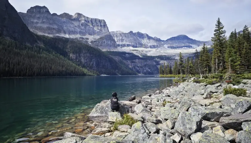 Persona sentada en rocas junto a un lago turquesa, contemplando majestuosas montañas nevadas. Un paisaje perfecto para viajar a Canadá.
