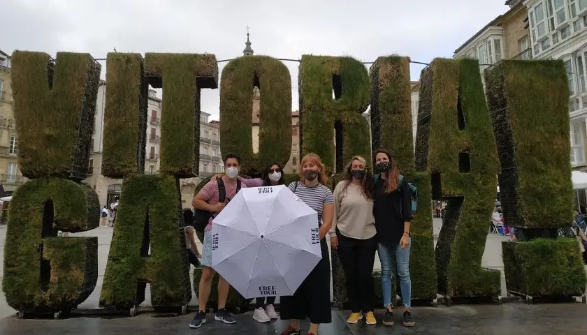 Grupo posa frente a letras gigantes de césped que dicen "VITORIA". Sostienen un paraguas blanco con el logo "FREE TOUR".